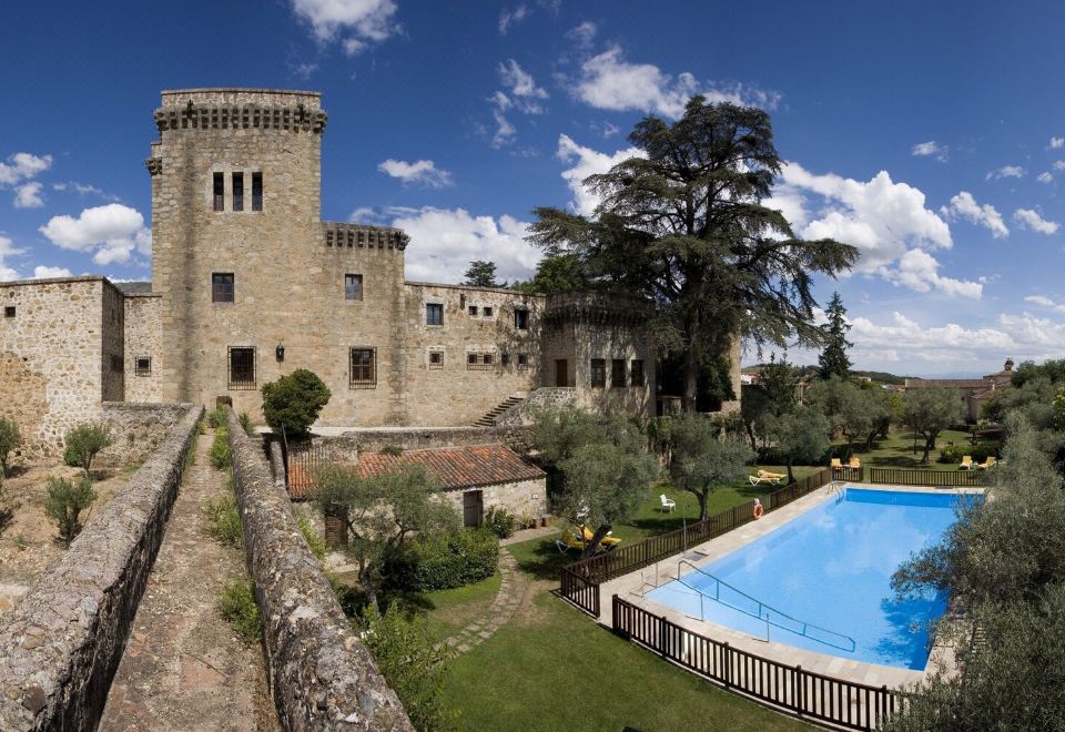 a large castle with a pool and trees in the foreground , under a blue sky with clouds at Parador de Jarandilla