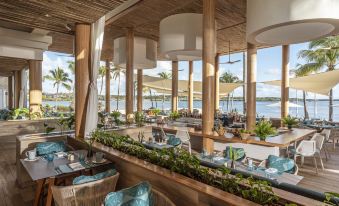 a restaurant with wooden tables and chairs , large windows , and tropical plants , overlooking the water at Four Seasons Resort Mauritius at Anahita