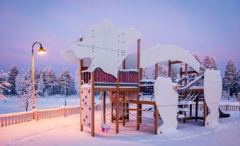 a snow - covered playground with a slide and a climbing structure , set against the backdrop of a winter landscape at Ranua Resort Holiday Villas