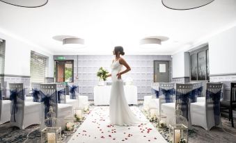 a woman in a white dress is standing in a room with flowers and white chairs at Birch Hotel