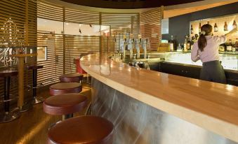 a modern bar with a curved wooden bar counter and several stools surrounding it , creating a cozy atmosphere at Novotel Leuven Centrum