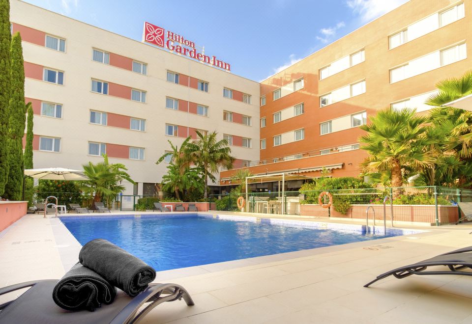 an outdoor pool surrounded by a hotel building , with a lounge chair and umbrellas placed around the pool area at Hilton Garden Inn Malaga