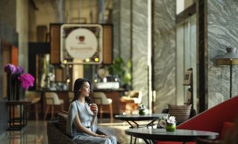 a woman in a blue dress is sitting on a chair in a hotel lobby , holding a cup of coffee at Swissôtel Jakarta Pik Avenue