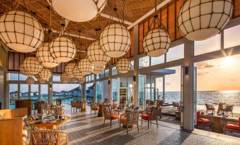 a large restaurant with wooden tables and chairs , hanging light fixtures , and a view of the ocean at Raffles Maldives Meradhoo Resort