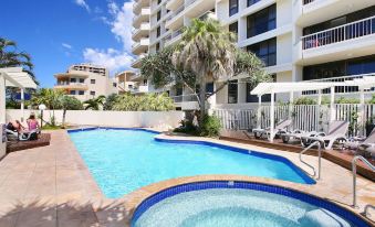 a large swimming pool is surrounded by a pool and lounge chairs in front of a tall white building at Coolum Caprice