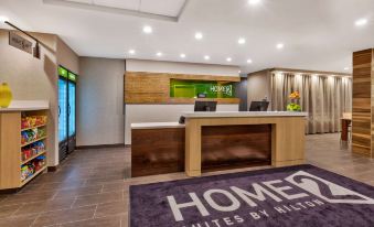 "a hotel lobby with a wooden reception desk , a green logo on the wall , and a sign for "" homewood suites by hilton "" in" at Home2 Suites by Hilton Battle Creek