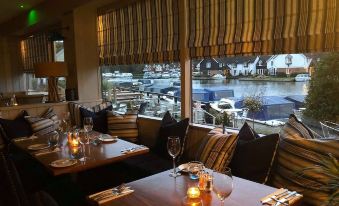 a restaurant with wooden tables , chairs , and wine glasses is shown with a view of a harbor at Hotel Wroxham