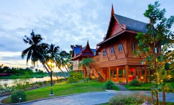 a wooden house with a red roof is surrounded by palm trees and is situated near a lake at RK Riverside Resort & Spa (Reon Kruewal)
