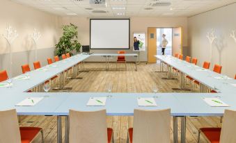 a large conference room with multiple long tables and chairs , a projector screen , and a plant at Ibis Mulhouse Bale Aeroport
