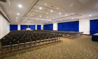 a large , empty auditorium with rows of black chairs and blue curtains , set against a white ceiling with exposed lights at Dann Carlton Medellin Hotel