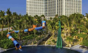 a large resort with a water park and a tall building in the background , surrounded by lush greenery at Movenpick Resort Cam Ranh