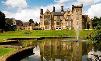 a large , stone building with multiple chimneys and a moat , surrounded by green grass and trees at Delta Hotels Breadsall Priory Country Club