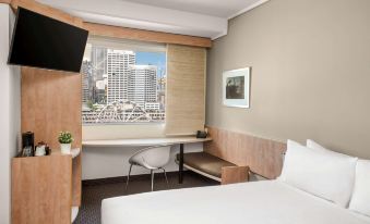 a modern hotel room with a large window offering a city view , wooden desk , and bed , as well as some plants and a small table at Ibis Sydney Darling Harbour