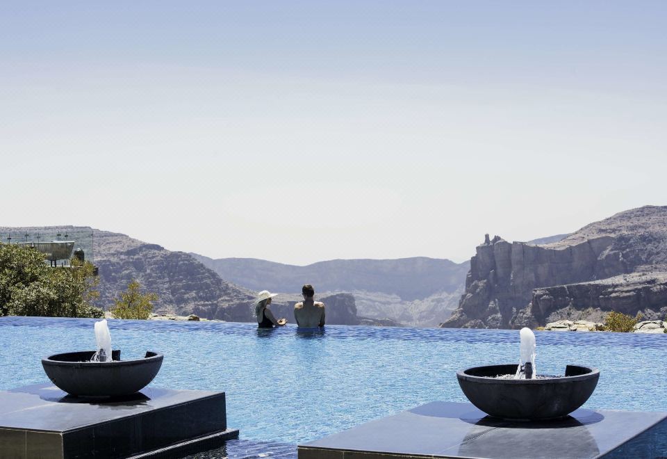 a man and a woman are standing in a large outdoor swimming pool , surrounded by mountains in the background at Anantara Al Jabal Al Akhdar Resort
