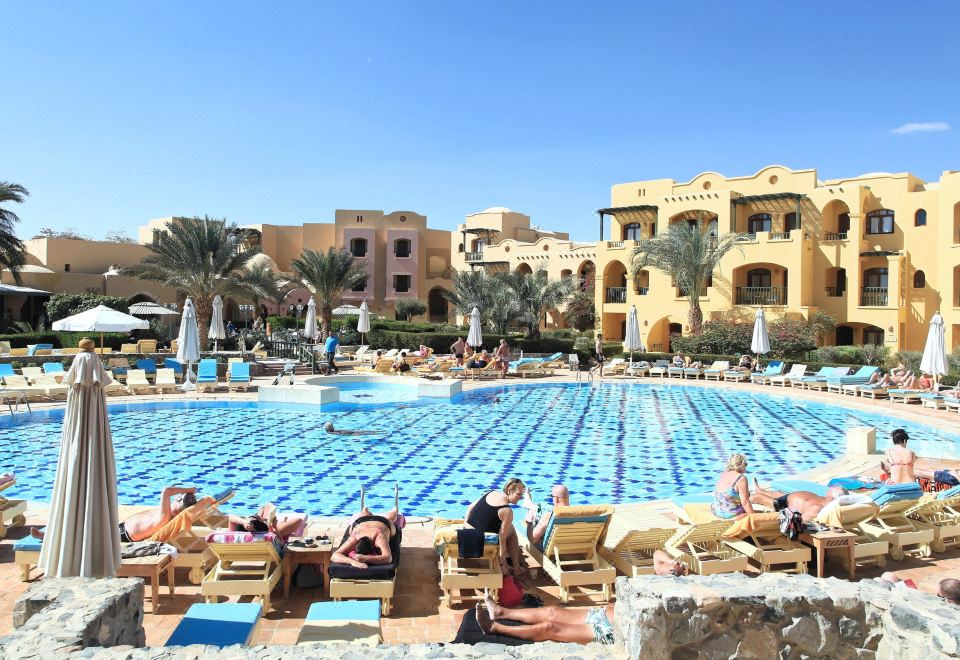 a large swimming pool with people in the water and lounge chairs surrounding it , near a building with palm trees at Three Corners Rihana Resort El Gouna