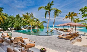 a large outdoor pool surrounded by lounge chairs and umbrellas , with palm trees in the background at Joali Maldives
