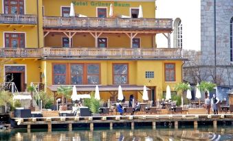 a large yellow building with a restaurant on the side , surrounded by trees and water at Seehotel Gruner Baum