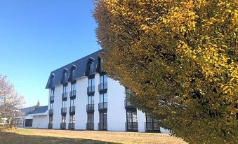 a white building with black windows is surrounded by a tree and yellow leaves , set against a clear blue sky at Gr8 Hotel Sevenum