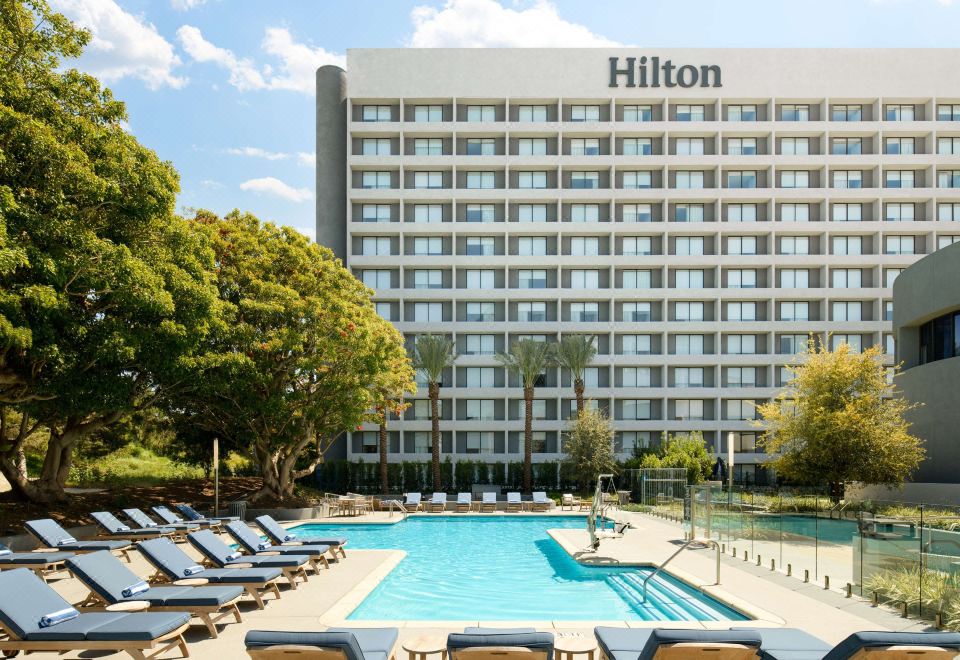 a large hotel with a swimming pool surrounded by lounge chairs and trees in front at Hilton Los Angeles-Culver City, CA