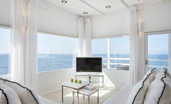 a modern living room with white walls , large windows offering views of the ocean , and a tv on a glass table at Notos Therme and Spa