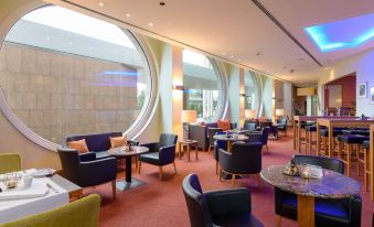 a modern lounge area with blue and black chairs , wooden tables , and large windows , providing a comfortable and inviting atmosphere at Novotel Freiburg am Konzerthaus