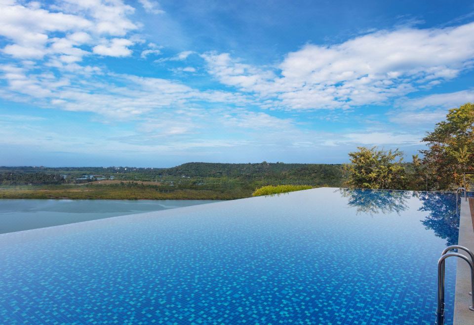 a large , blue infinity pool overlooking a grassy field and a body of water under a cloudy sky at DoubleTree by Hilton Goa - Panaji