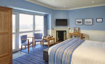 a hotel room with a king - sized bed , a fireplace , and a large window overlooking the ocean at Trearddur Bay Hotel
