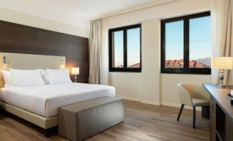 a hotel room with a white bed , wooden floor , and two large windows overlooking a city view at Doubletree by Hilton Brescia
