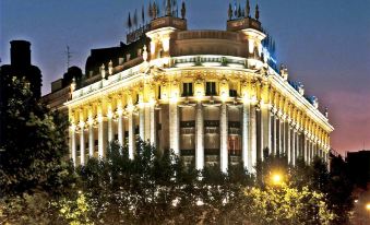 a large building with a white facade and columns is lit up at night , surrounded by trees and fountains at NH Madrid Nacional