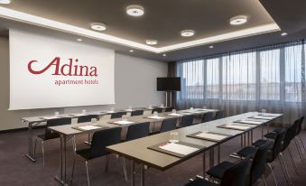 a conference room with multiple tables and chairs , a large screen displaying the adina logo , and large windows at Adina Apartment Hotel Nuremberg