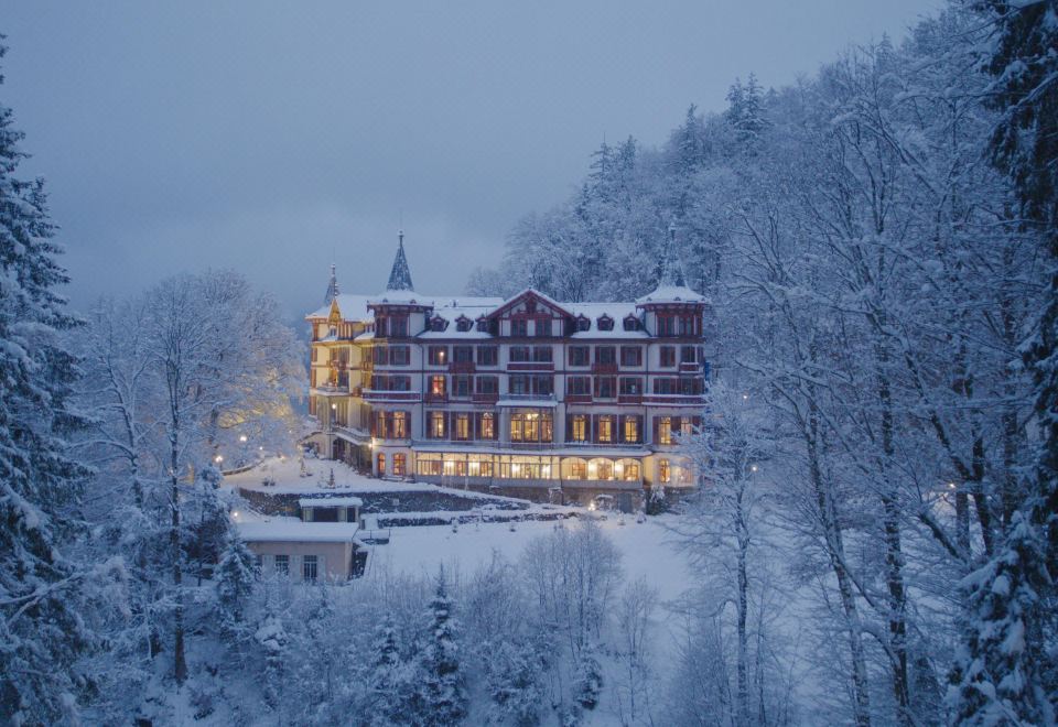 a large , red - roofed hotel surrounded by snow - covered trees and mountains , with lights illuminating the building at Grandhotel Giessbach