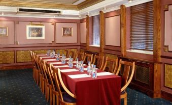 a long dining table with red tablecloths and wooden chairs , set up in a room with pink walls and windows at Berjaya Eden Park London Hotel