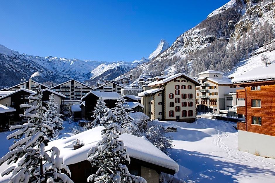 a snow - covered village surrounded by mountains , with a few buildings and trees covered in snow at Hotel Ambassador Zermatt