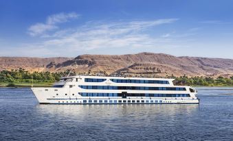 a large cruise ship docked in a body of water , with mountains in the background at The Oberoi Zahra, Luxury Nile Cruiser
