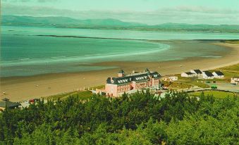 a large , pink building is situated on the shore of a beach with mountains in the background at Sandhouse Hotel