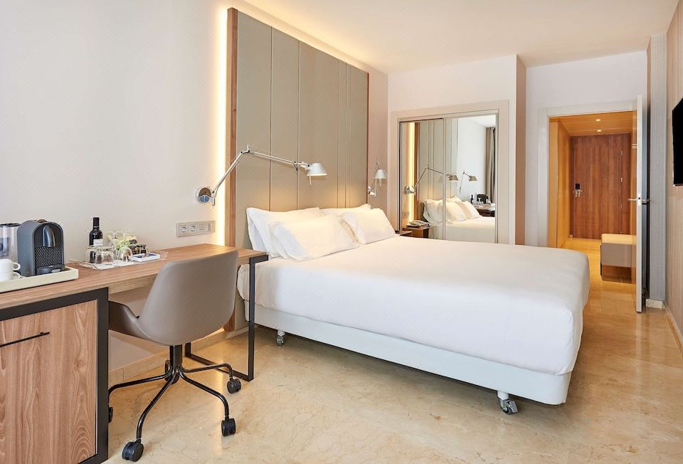 The spacious bedroom features a double bed, a desk, and a chair, all set against the backdrop of white walls at NH Logrono Herencia Rioja