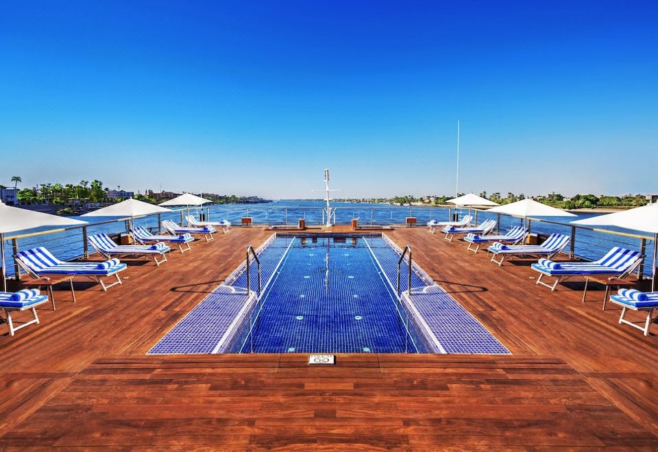 a large swimming pool surrounded by wooden decking , with lounge chairs and umbrellas placed around it at The Oberoi Zahra, Luxury Nile Cruiser