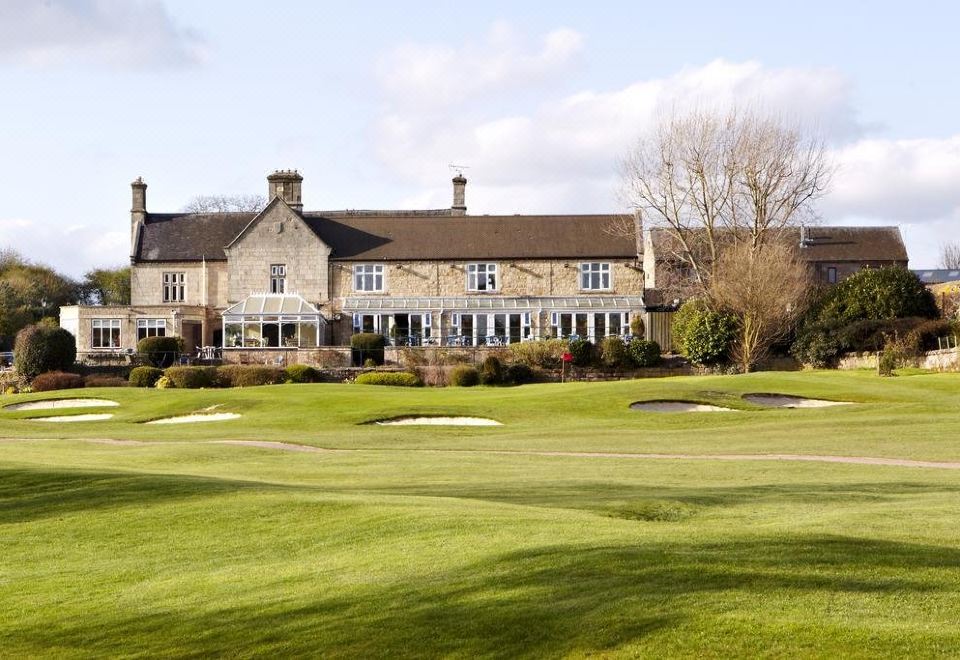 a large stone building with a sloping roof is situated on a golf course , surrounded by trees and grass at Horsley Lodge