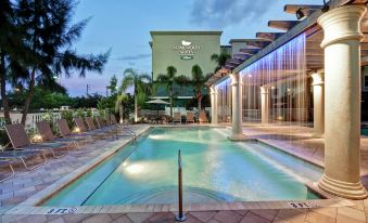 a large outdoor swimming pool surrounded by lounge chairs and umbrellas , with a building in the background at Homewood Suites by Hilton Tampa-Port Richey