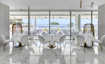 a modern , white - themed restaurant with large windows offering a view of the sea , featuring several white chairs and tables set up for dining at The Ivi Mare - Designed for Adults by Louis Hotels