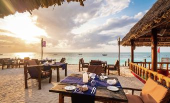 a beachside restaurant with wooden tables and chairs , where people are enjoying their meals and the view of the ocean at Nungwi Beach Resort by Turaco