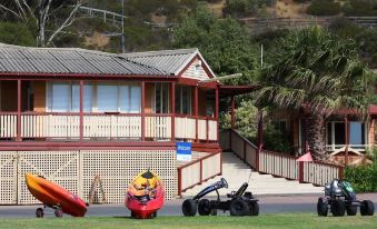 a wooden building with a red roof , surrounded by trees and mountains , and two people riding a lawn mower in front of it at Brighton Beachfront Holiday Park Adelaide