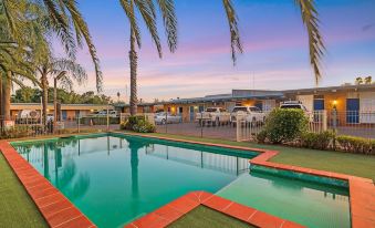 a large swimming pool surrounded by palm trees , with a building in the background and cars parked in front at Plainsman Motel