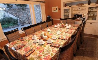 a large dining table is set with plates , cups , and a floral tablecloth in a room with windows at Arroyo Vista Inn