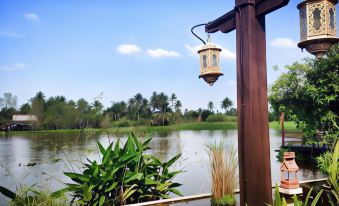 a serene scene of a lake surrounded by trees and a wooden fence , with a lantern hanging from a pole in the foreground at Parn Dhevi Riverside Resort & Spa