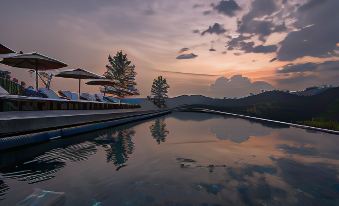 a large outdoor pool with lounge chairs and umbrellas , surrounded by trees and mountains , at sunset at Santani Wellness Kandy