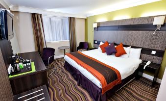 a hotel room with a king - sized bed , a chair , and a tv . the room has a striped carpet , purple chairs , and at Link Hotel