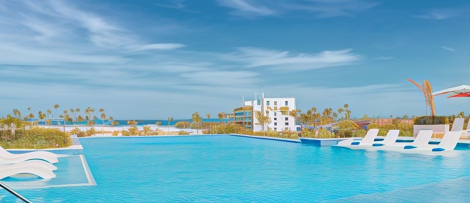 a large blue pool is surrounded by a white building and palm trees , with a clear blue sky in the background at Radisson Blu Aruba
