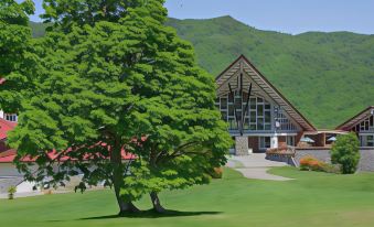 a large tree is in front of a building with mountains and green grass in the background at Okushiga Kogen Hotel