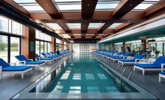 a large indoor swimming pool with blue lounge chairs and a glass ceiling , surrounded by glass windows at Burgu Arjaan by Rotana